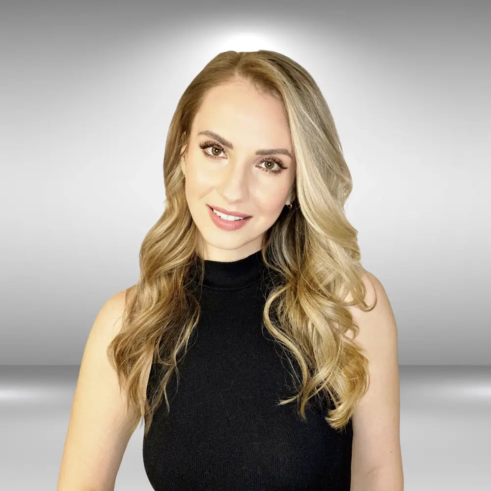 A talented woman with long, wavy, blonde hair and light skin smiles at the camera, exuding confidence. She is wearing a sleeveless black top against a light grey background with a subtle gradient effect, ready for her audition.