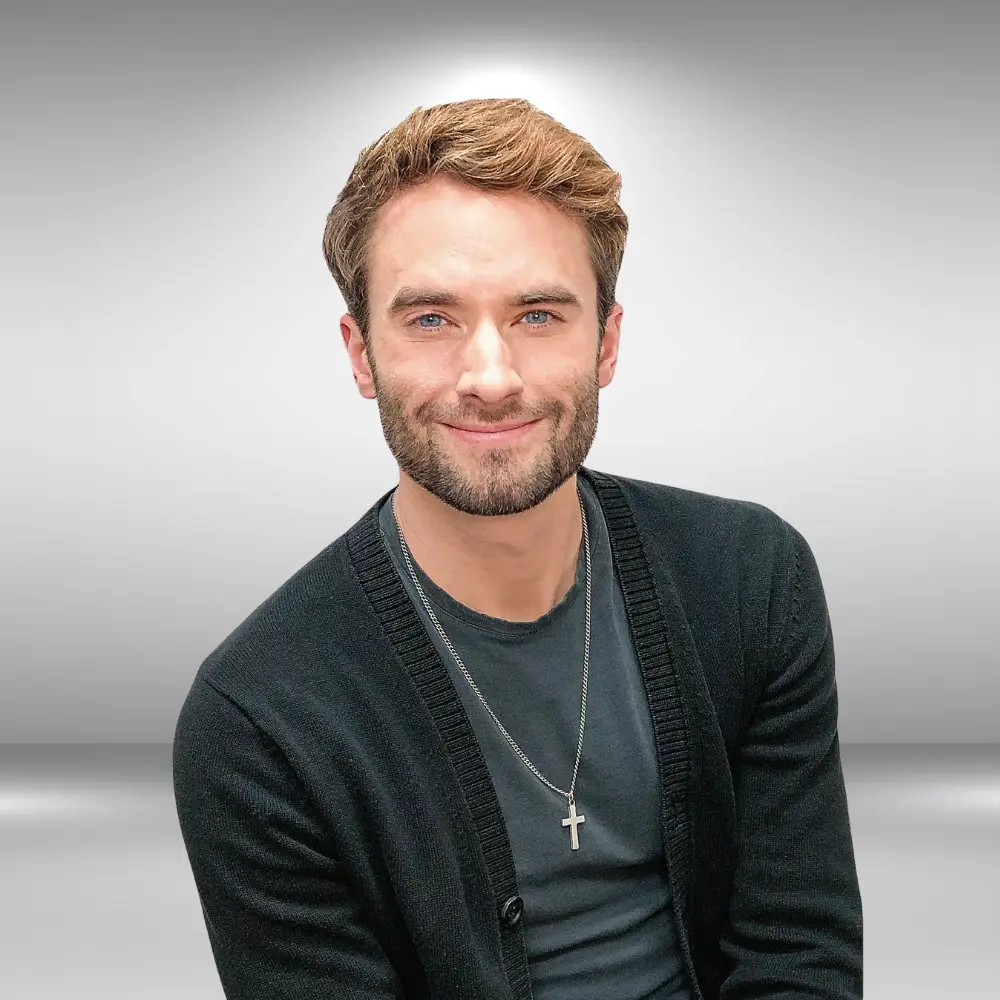 A man with short brown hair and a beard is smiling at the camera. He is wearing a black cardigan over a dark shirt, and a silver cross necklace. The background is a plain gradient from light gray at the top to darker gray at the bottom, perfect for his audition profile as an up-and-coming actor.