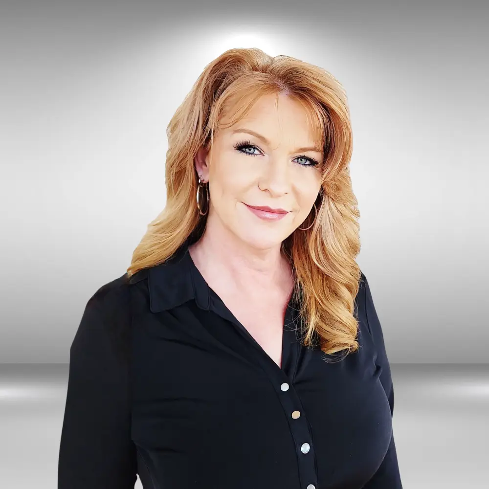 A woman with wavy, shoulder-length ginger hair and blue eyes is smiling gently. She is wearing a black button-up shirt with hoop earrings and is standing against a grey gradient background, exuding the confidence of a seasoned influencer ready for her next big audition.