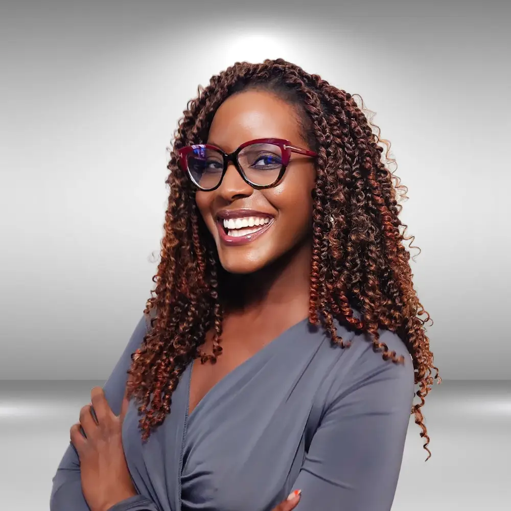 A talented woman with long curly hair, wearing glasses and a grey blouse, smiles brightly with her arms crossed. She stands against a grey gradient background.