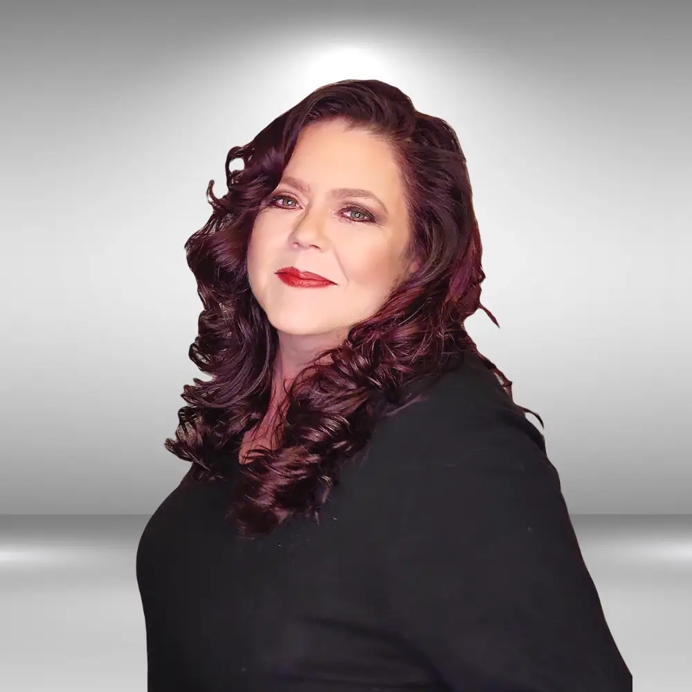 A woman with long, wavy dark hair and red lipstick is posing against a light gray gradient background. She is wearing a black top and is smiling slightly, looking directly at the camera. Her head is tilted slightly to the side, showcasing her talent as an influencer.