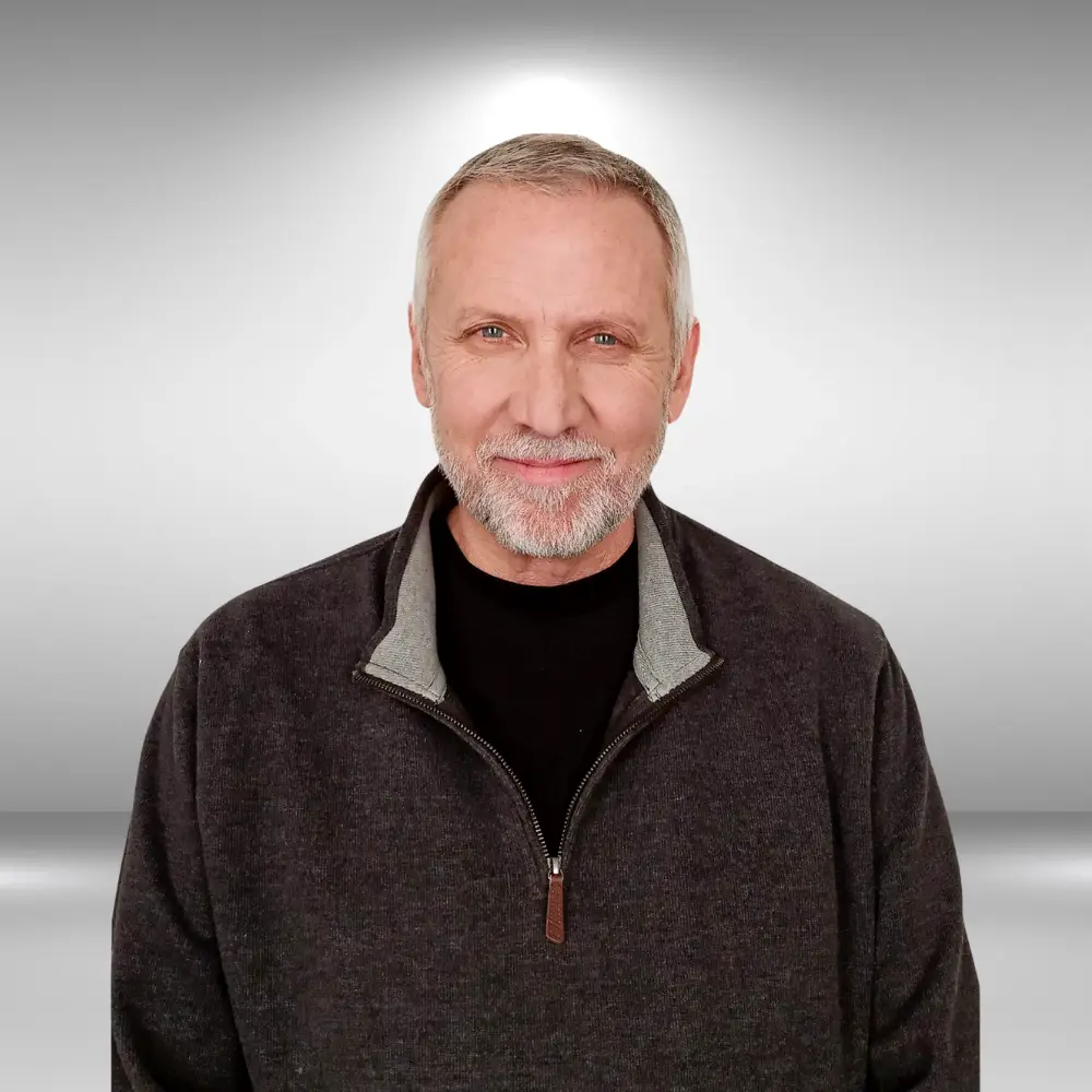 A smiling man with short gray hair and a beard, wearing a dark gray zip-up sweater over a black shirt, stands against a plain gray background, exuding talent and confidence as if ready for an audition or to walk the runway.