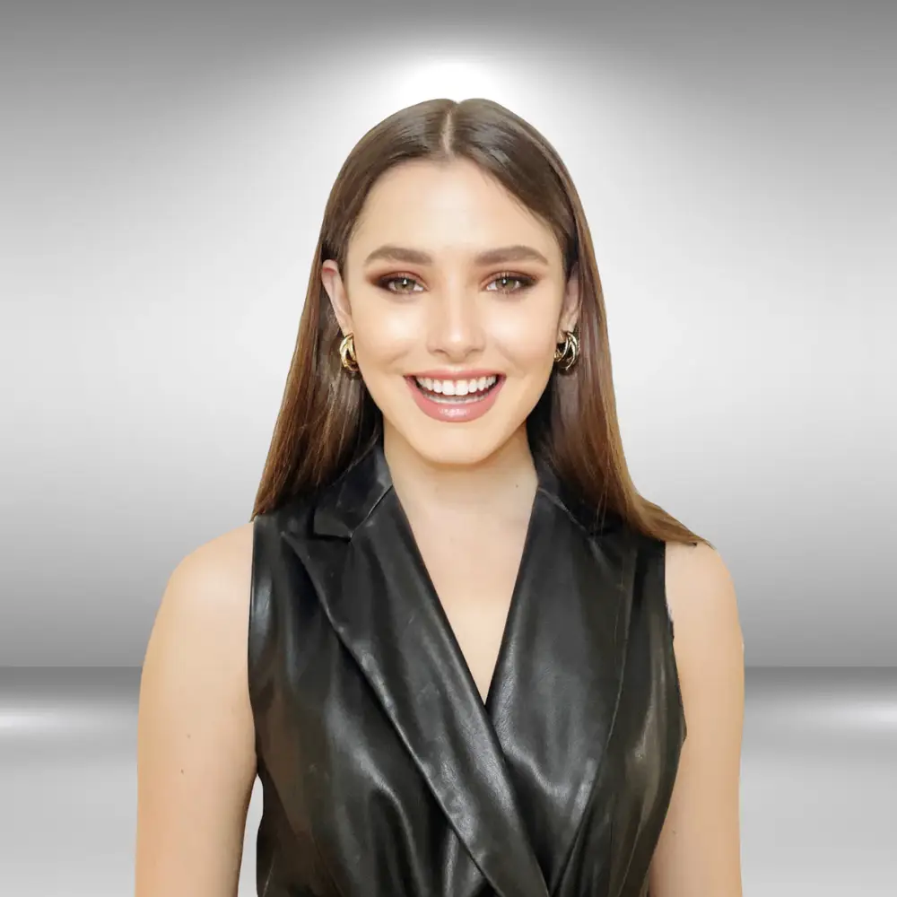 A woman with long brown hair is smiling warmly. She is wearing a sleeveless black leather top or dress, and gold hoop earrings. The background is a gradient of light gray to white, creating a soft halo effect around her head, giving her the perfect look for an audition.